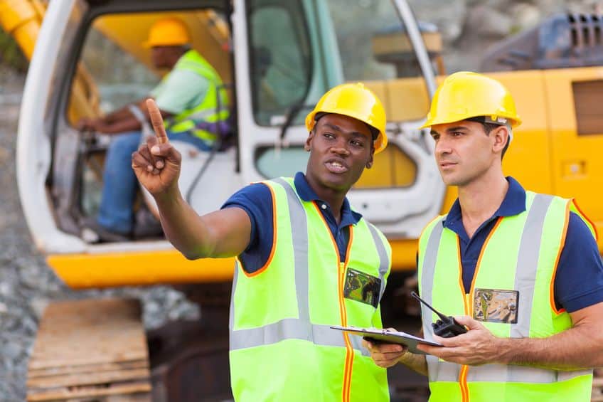 two construction workers talking a construction site with a coworker operating a bulldozer behind them