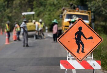 under construction symbol on an orange sign with construction workers working on the road in the background