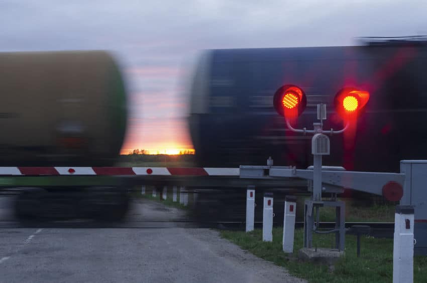 railroad lights lit up with a barrier down and a train behind it moving quickly on the tracks, a sunset is in the background