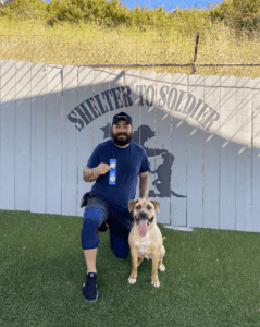 man with dog standing in front of a fence with the shelter to soldier logo on it